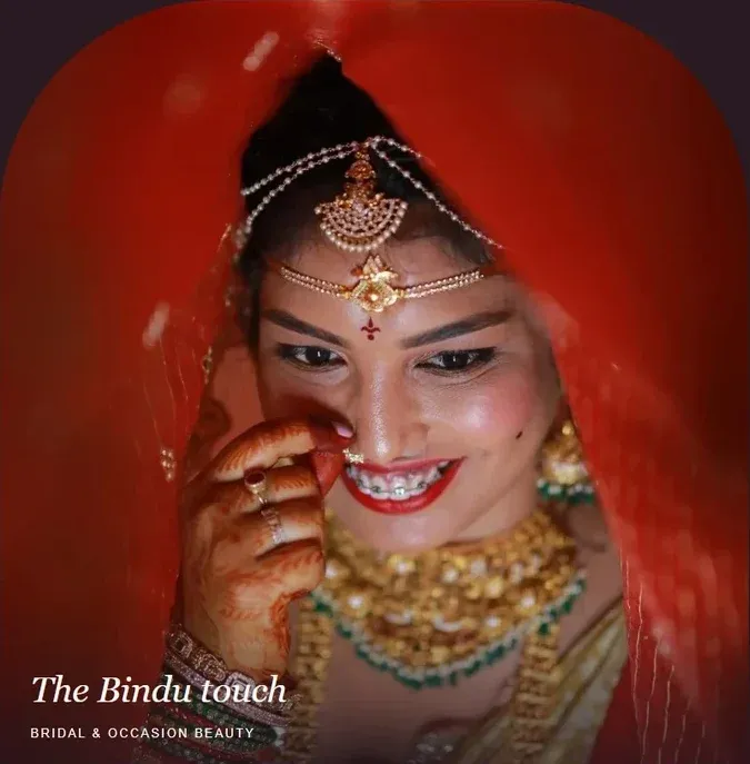 A smiling bride in a red dupatta with a maang tikka, nose ring and gold necklaces, one hand with mehndi raised to her face.